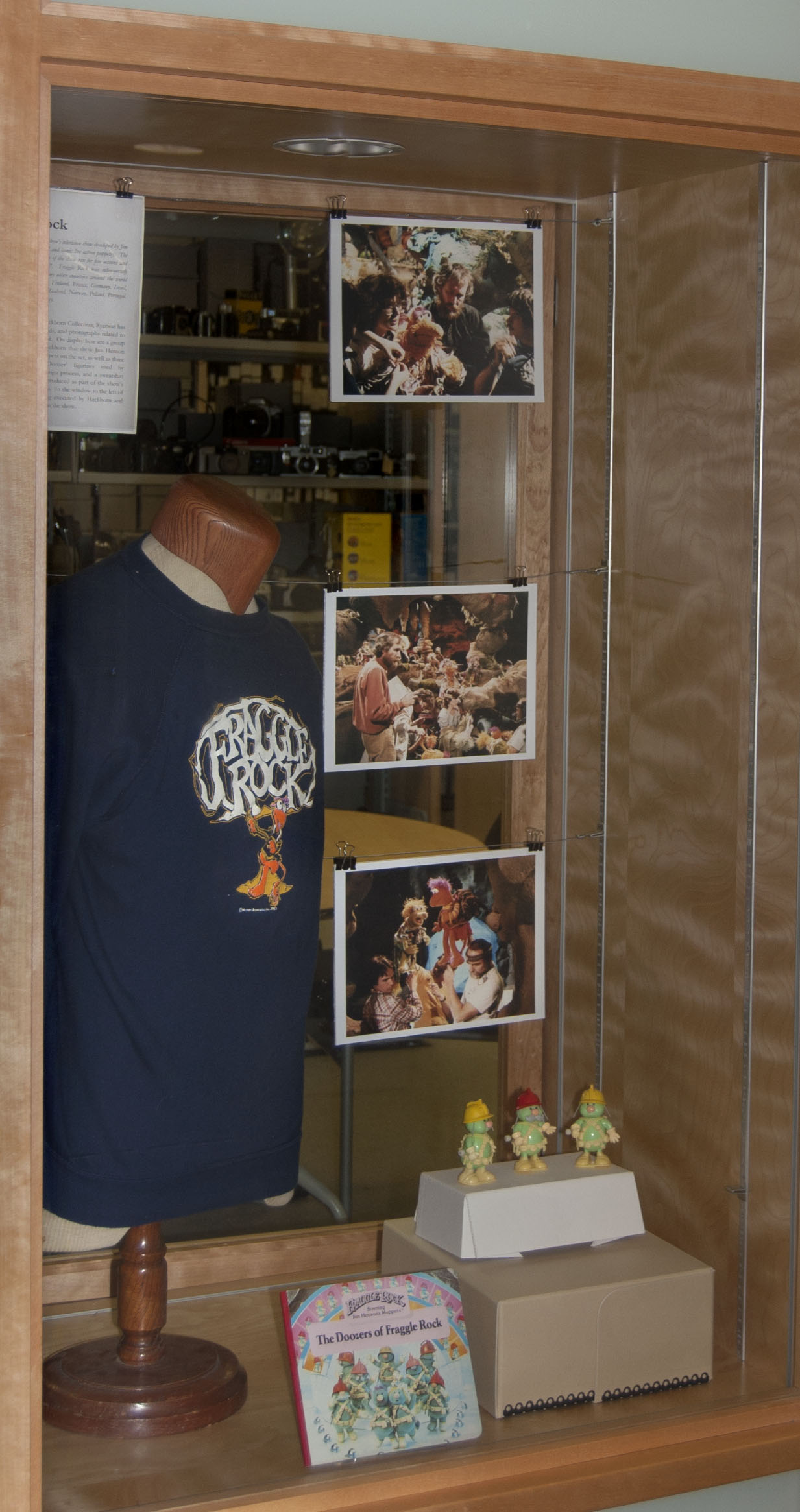 On display are a group of color photographs taken by Hackborn, a sweatshirt, a children’s picture book, and figurines related to the production of Fraggle Rock.