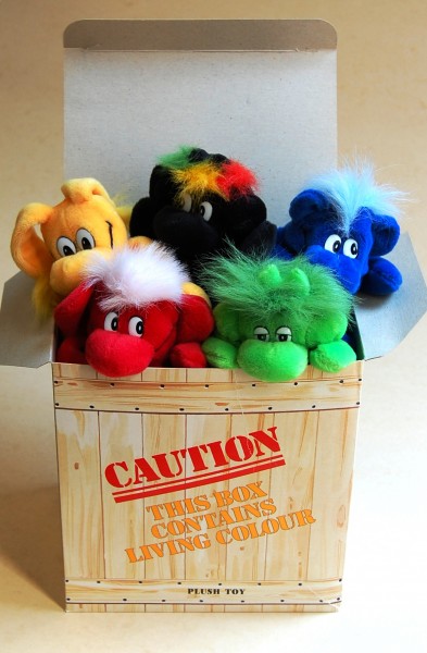 5 small plush toys (yellow, black, blue, green and red) in a cardboard box that reads "Caution this box contains living color"