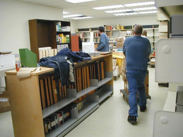 Office with many shelves containing albums, books and archival boxes. People moving boxes on carts. 