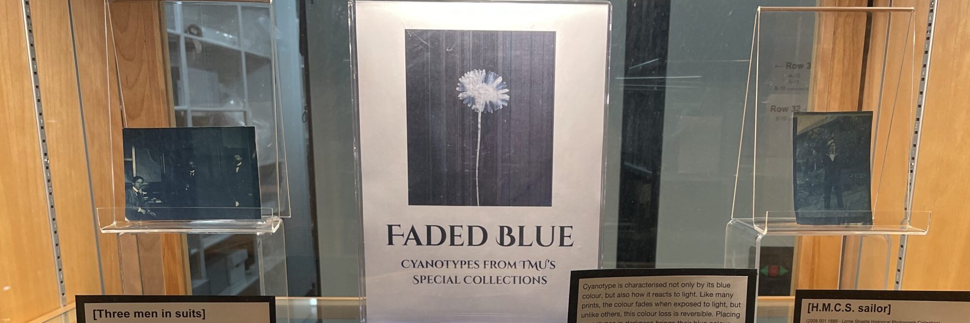 Interior of a display case. Shelf has a white and blue letter sized label, and two blue photographs on risers. 