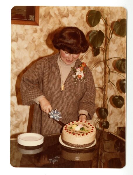 Woman in a brown jacket with a flower pin cutting a round cake with red and pink frosting.