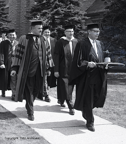 Line of men in black robes and mortarboards leaving building along tree lined path. Man in front is carrying an oil lamp on a pillow