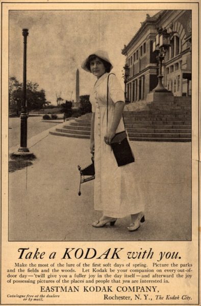 Image of a 1912 advertisement for Kodak Cameras, featuring a photograph of a women in a white dress, carrying a Kodak camera and case, and the slogan "Take a Kodak With You!".