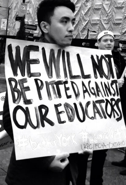 Black and white image of a person holding a sign with the text "We will not be pitted against our educators #BetterYork #WeareUofT" painted in black letters.